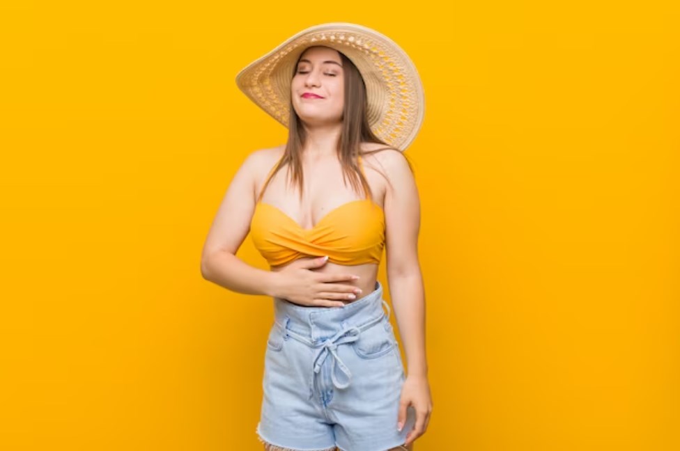 a young smiling caucasian woman wearing a straw hat and touching her tummy and representing satisfaction