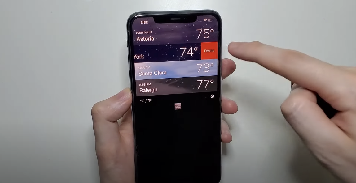 A picture of a hand holding a phone displaying the weather of three cities, with an attempt to delete one of the cities